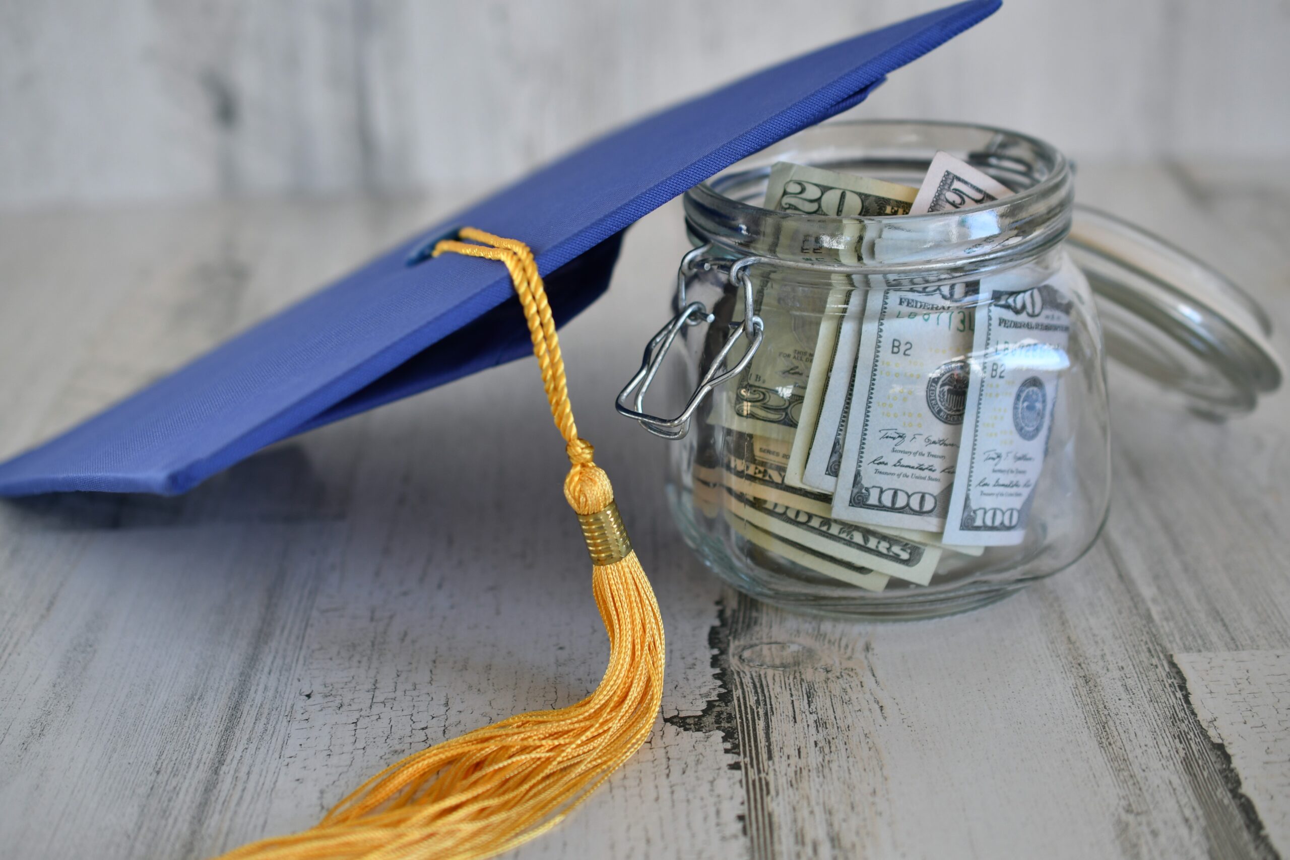 Graduation cap mortarboard with tassel next to jar of saved money cash.